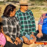 A group of Aboriginal and Torres Strait Island people are sitting in the Australian bush looking towards a Diabetes Australia First Nations health worker while talking.