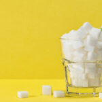 Against a bright yellow background, a glass with white sugar cubes has a blue and white straw sticking out of it, with sugar cubes lying around its base.