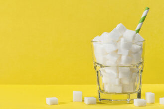 Against a bright yellow background, a glass with white sugar cubes has a blue and white straw sticking out of it, with sugar cubes lying around its base.