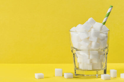 Against a bright yellow background, a glass with white sugar cubes has a blue and white straw sticking out of it, with sugar cubes lying around its base.