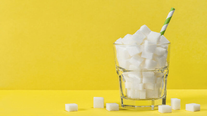 Against a bright yellow background, a glass with white sugar cubes has a blue and white straw sticking out of it, with sugar cubes lying around its base.