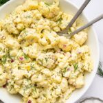 Cauliflower potato salad in a large serving bowl with silver serving utensils. The salad is topped with fresh dill.