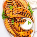 Grilled sweet potatoes on a serving platter topped with fresh parsley and served with a side of garlic aioli. There is one sweet potato dipped in the aioli.