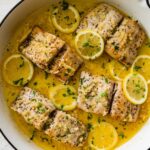4 Mahi mahi fillets in a skillet with a lemon butter garlic sauce. There are lemon slices in the skillet and everything is topped with fresh parsley.