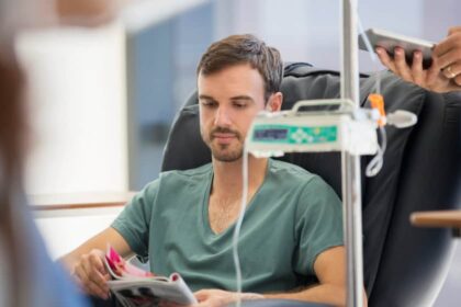 A young man sits in a hospital chair reading a magazine with drips and an iv in the frame.