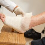 A foot propped on a wooden stool is carefully bandaged by a health professional wearing gloves.