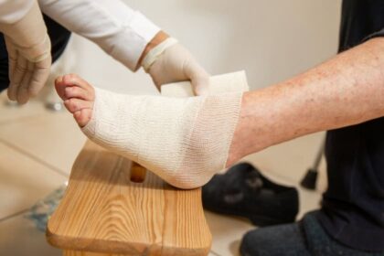 A foot propped on a wooden stool is carefully bandaged by a health professional wearing gloves.