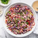 Assembled Asian slaw in a white bowl, garnished with sesame seeds and scallions. Dressing and toppings sit around the bowl on a marble counter with a striped towel.