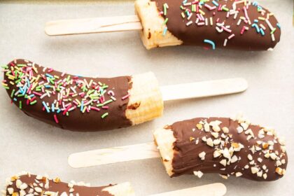 Frozen banana halves on wooden sticks, dipped in chocolate and topped with various toppings including rainbow sprinkles, chopped nuts, and shredded coconut.