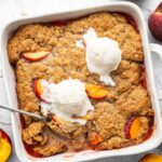 Freshly baked healthy peach cobbler in a white baking dish topped with vanilla ice cream and a spoon scooping out a serving.