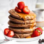 Tall stack of chocolate protein pancakes topped with fresh strawberries and nut butter drizzle.